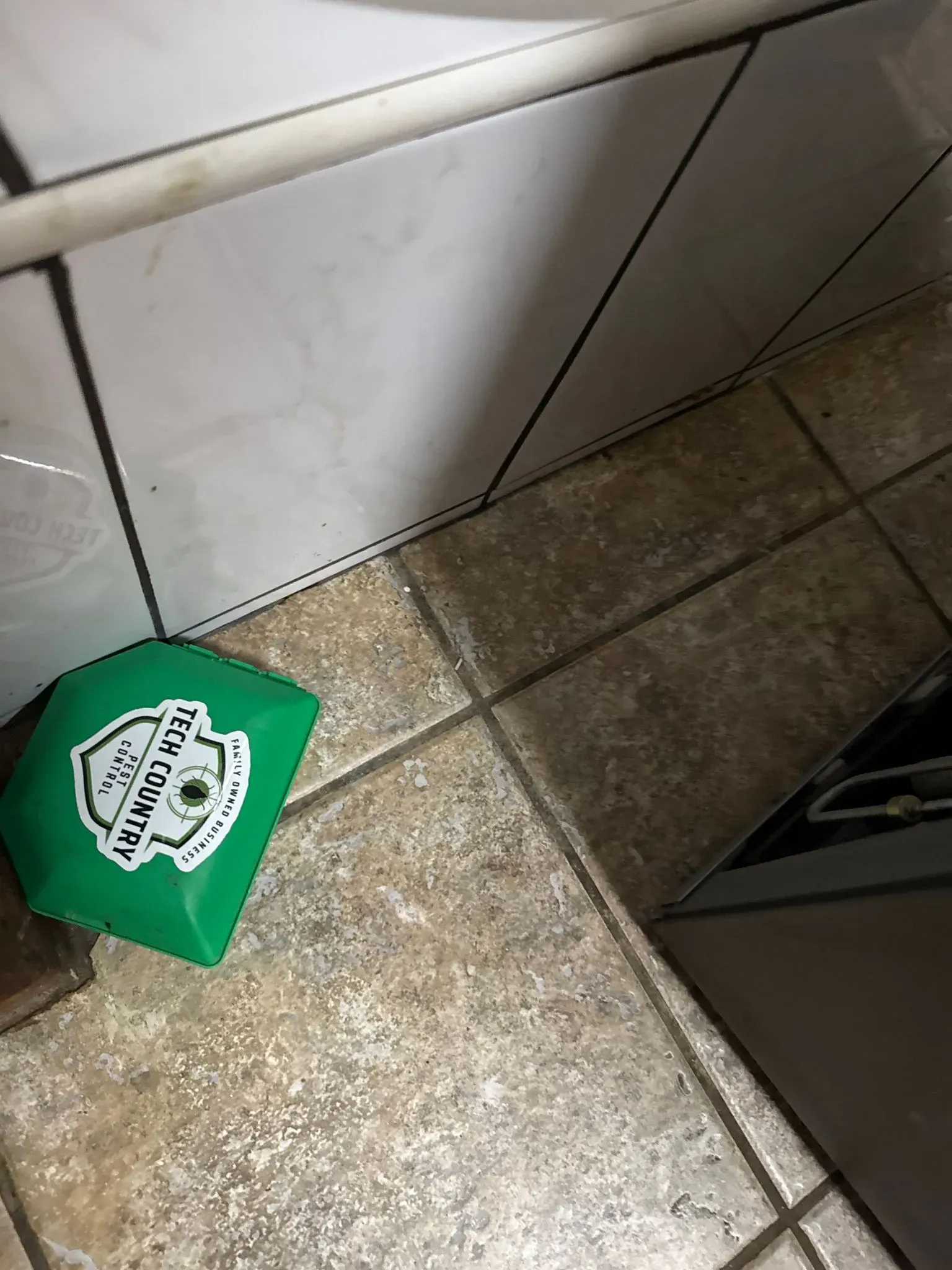 A green pest control bait station with the Tech Country Pest Control logo on a tiled floor.