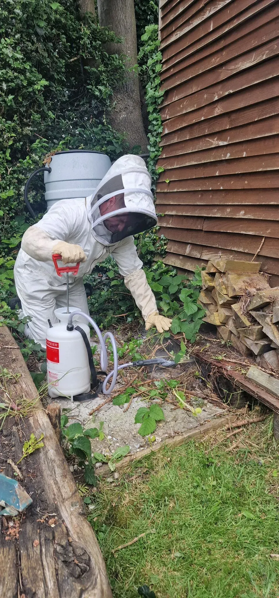 A pest control technician in a white protective suit and hood, kneeling and using a sprayer to treat an area on the ground next to a brown wooden shed, surrounded by green foliage.